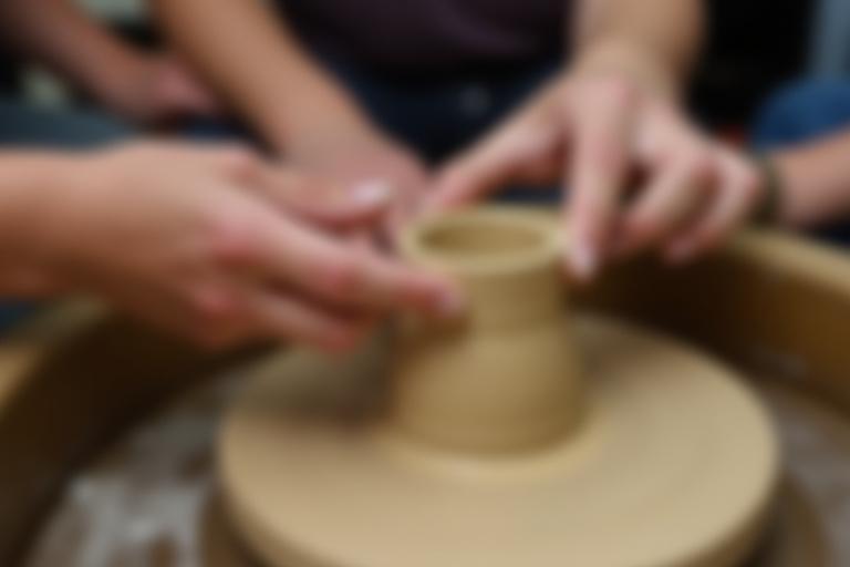 Eine Nahaufnahme von Händen, die auf einer Töpferscheibe Ton formen, während eines Keramikkurses.